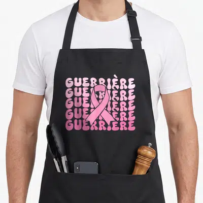 Tablier de cuisine noir avec le mot GUERRIÈRE en rose et un ruban symbolique au centre.