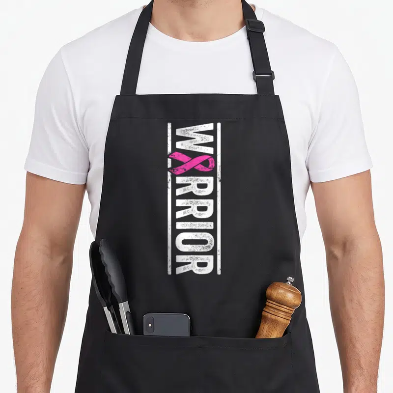 Tablier de cuisine noir avec l'inscription verticale Warrior et un ruban rose stylisé au centre du design.