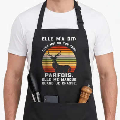 Tablier de cuisine noir avec un cerf gris devant un soleil rayé orange vert et texte blanc humoristique sur la chasse.