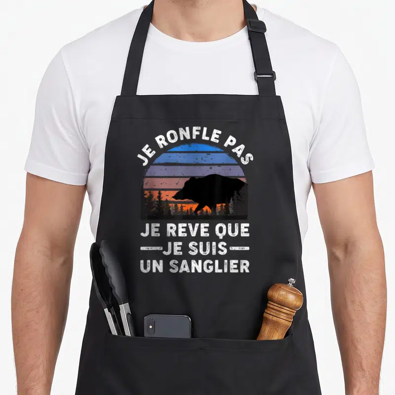 Tablier de cuisine noir avec silhouette de sanglier et texte blanc sur fond de soleil couchant coloré dans la forêt.