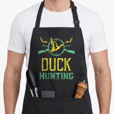 Tablier de cuisine noir avec illustration jaune et verte d'un canard en vol sur fond de viseur et fusils croisés.