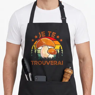 Tablier de cuisine noir avec illustration vintage de cèpes et texte Je Te Trouverai sur fond de soleil couchant.