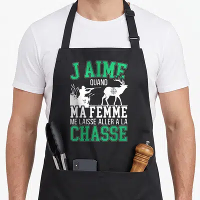 Tablier de cuisine noir avec inscription verte et blanche J'aime quand ma femme me laisse aller à la chasse sur fond gris uni.