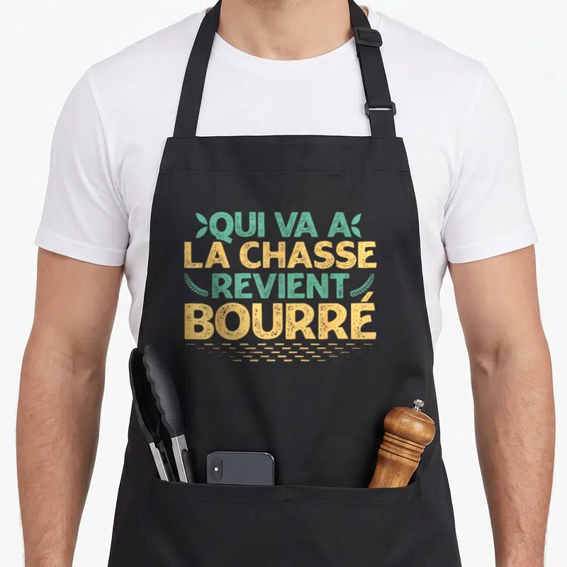 Tablier de cuisine noir avec le texte « Qui va à la chasse revient bourré » en typographie colorée et motifs végétaux stylisés.