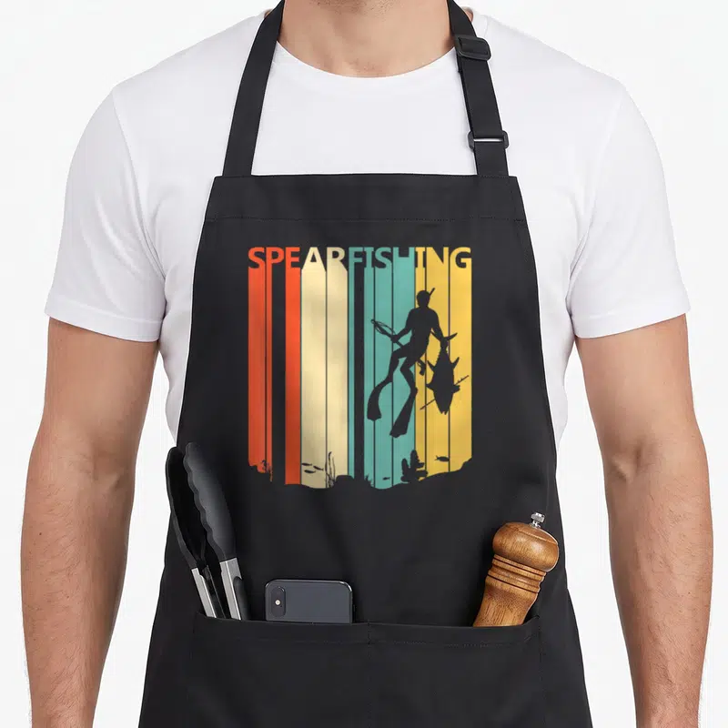 Tablier de cuisine noir avec illustration rétro d'un plongeur en apnée tenant une lance à poisson devant des bandes colorées.