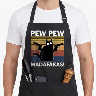 Tablier de cuisine noir avec un chat noir tenant deux pistolets et le texte Pew Pew Madafakas en blanc.