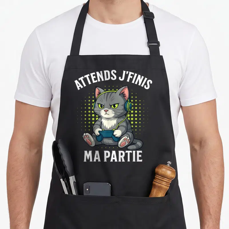 Tablier de cuisine noir avec un chat gris aux écouteurs tenant une manette de jeu vidéo et le texte Attends j'finis ma partie.