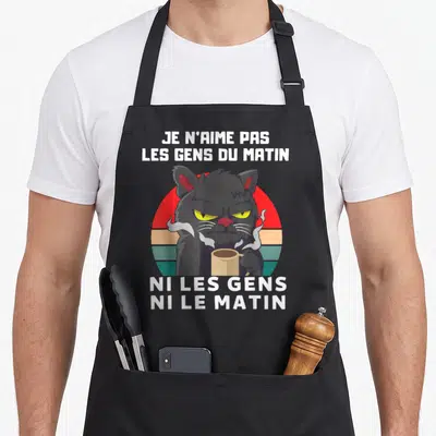 Tablier de cuisine noir avec un chat gris grincheux tenant une tasse de café sur fond rétro coloré.