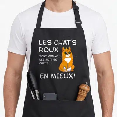 Tablier de cuisine noir avec un chat roux coiffé d'une couronne et un texte humoristique en blanc.
