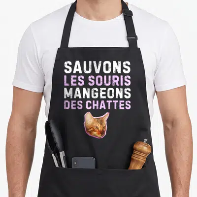 Tablier de cuisine noir avec texte blanc et rose Sauvons les souris mangeons des chattes au-dessus d'une tête de chat tigré.