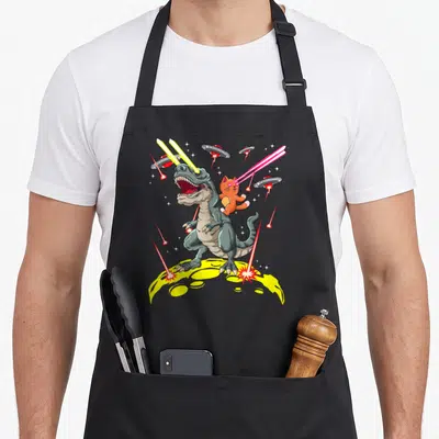 Tablier de cuisine noir avec illustration d'un chat orange sur un dinosaure spatial émettant des lasers jaunes et roses.
