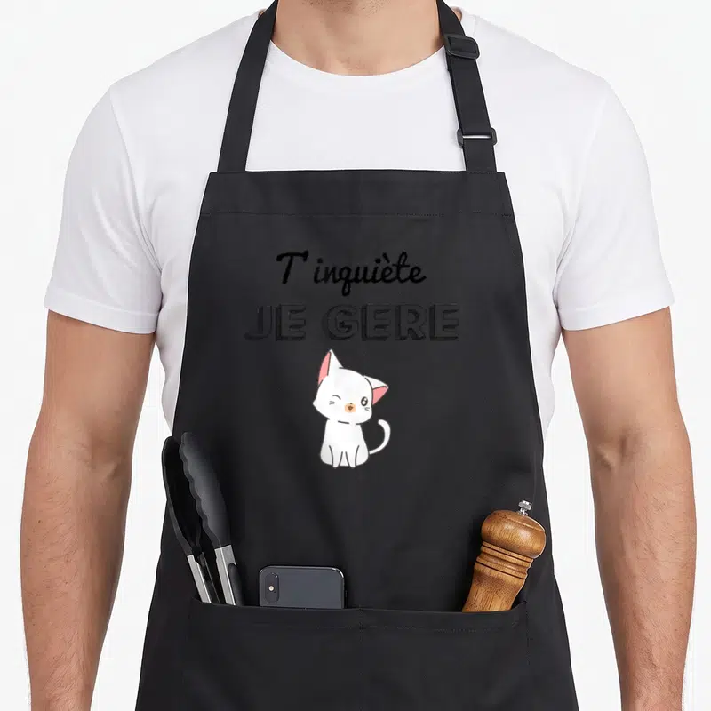 Tablier de cuisine noir avec un chat blanc kawaii et l'inscription T'inquiète Je Gère en typographie noire.
