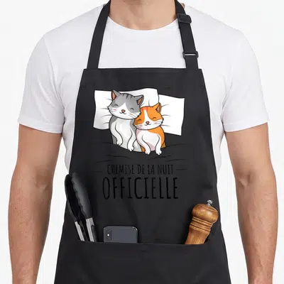 Tablier de cuisine noir avec deux chats blottis sur un oreiller blanc et l'inscription « chemise de la nuit » en lettres noires.