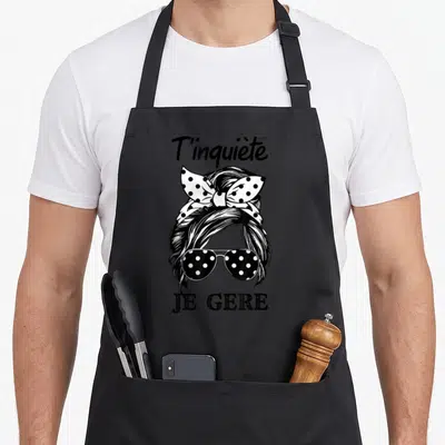 Tablier de cuisine noir avec illustration femme chignon, lunettes à pois et phrase T'inquiète Je Gère en typographie noire.