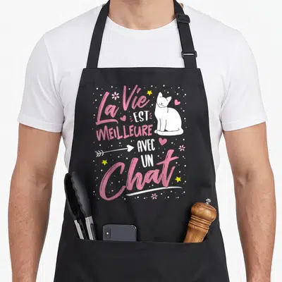 Tablier de cuisine noir avec inscription La Vie est meilleure avec un Chat et dessin de félin blanc entouré d'étoiles jaunes et de cœurs roses.