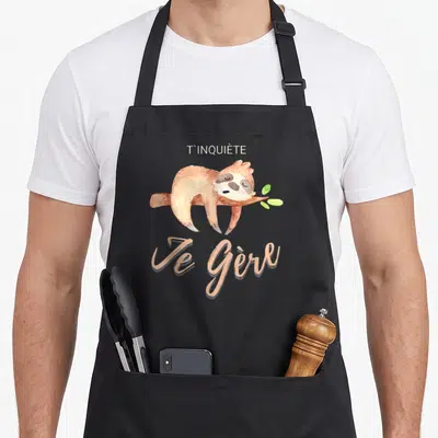 Tablier de cuisine noir avec un fourmis dormant sur une branche et le texte T'inquiète Je Gère imprimé en beige.