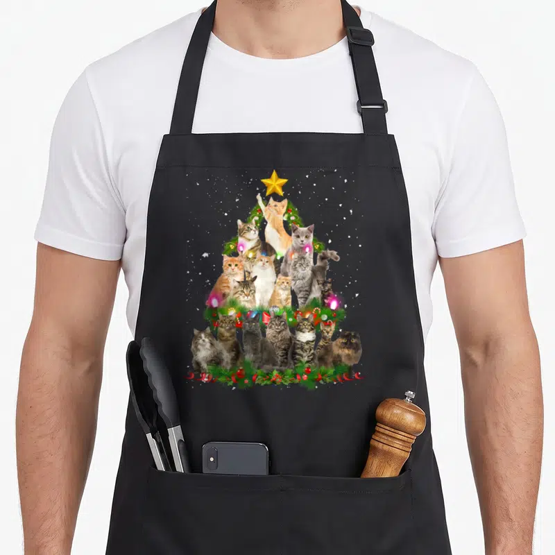 Tablier de cuisine noir avec une illustration d'arbres de Noël composés de chats et de décorations festives colorées.