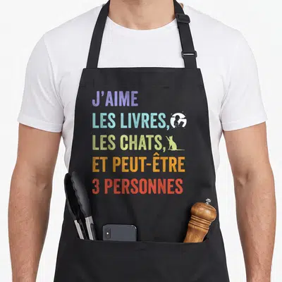 Tablier de cuisine noir avec inscription multicolore J'aime les livres les chats et peut-être 3 personnes accompagnée d'icônes stylisées.