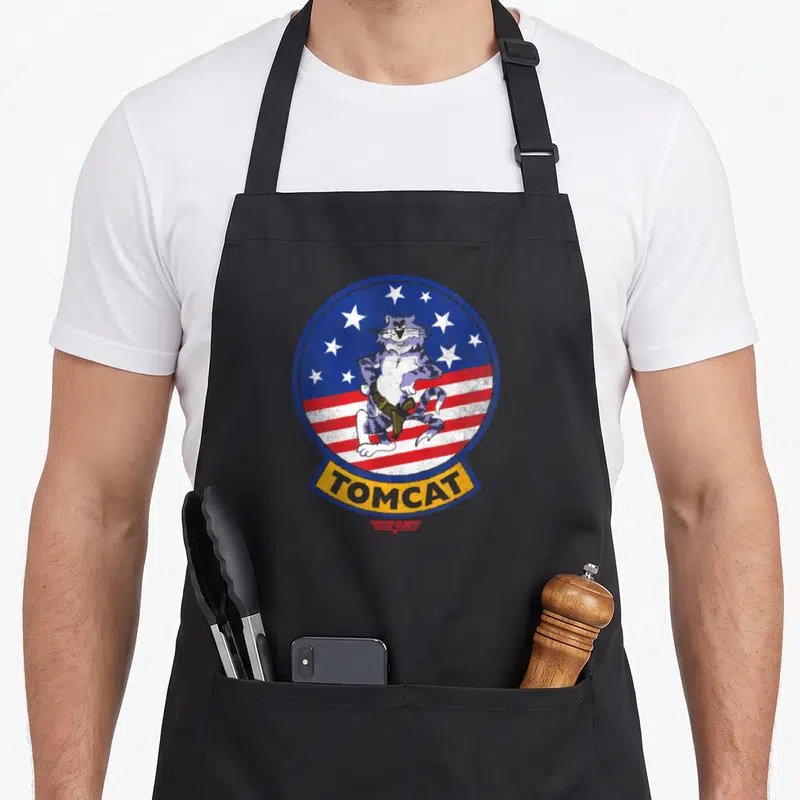 Tablier de cuisine noir avec un chat tigré en tenue de pilote sur fond d'étoiles et bandes rouges, inscription Tomcat en bas.
