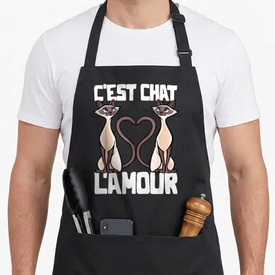 Tablier de cuisine noir avec deux chats aux tails entrelacés en cœur et le texte C'est Chat l'Amour en blanc.