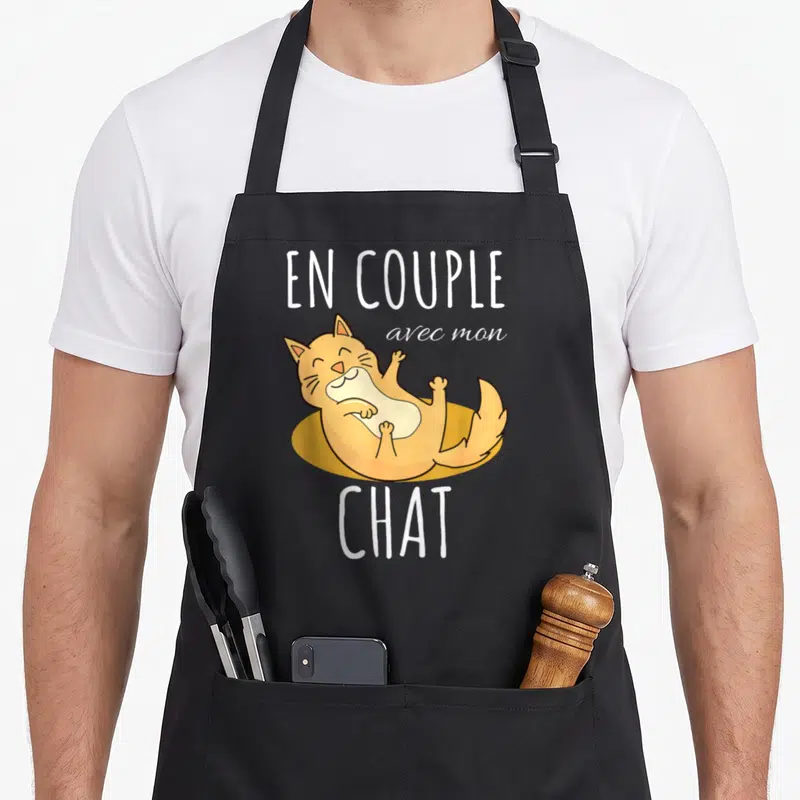 Tablier de cuisine noir avec un chaton orange allongé sur le dos et le texte En couple avec mon chat en blanc.