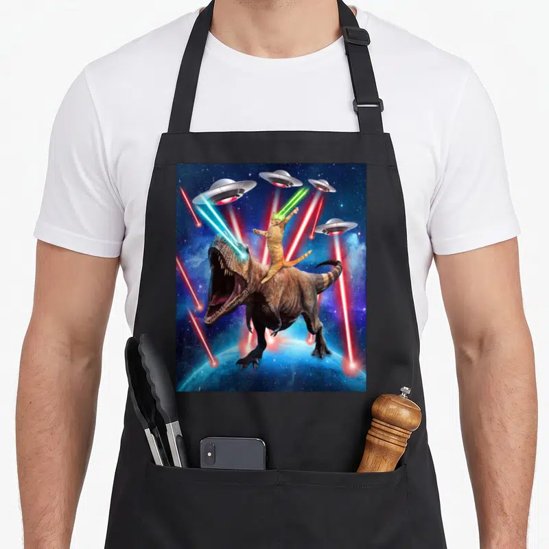 Tablier de cuisine noir avec illustration d'un chat tigré monté sur un dinosaure T-Rex sous des Ufo tirant des lasers spatiaux.