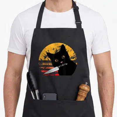 Tablier de cuisine noir avec illustration de chat noir tenant un couteau devant un soleil vintage orange et rouge.