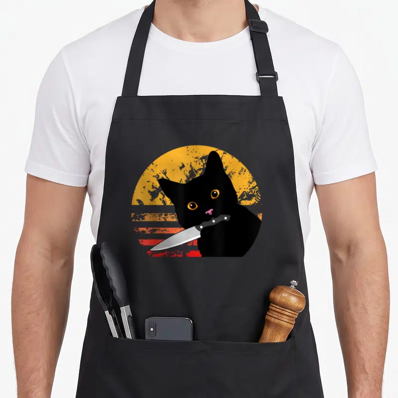 Tablier de cuisine noir avec illustration de chat noir tenant un couteau devant un soleil vintage orange et rouge.