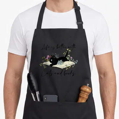 Tablier de cuisine noir avec un chat noir allongé sur un livre ouvert entouré de fleurs colorées et d'une inscription en anglais.