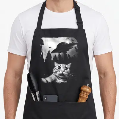 Tablier de cuisine noir avec illustration d'un chat taking a selfie sous des engins volants et des arbres stylisés.