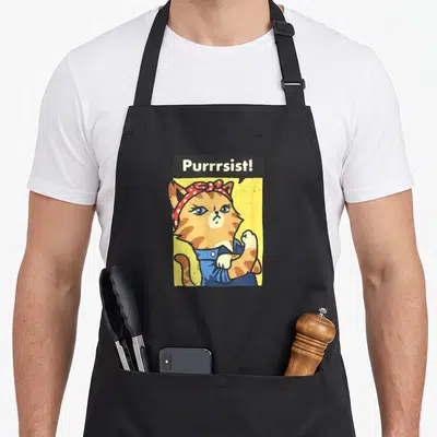 Tablier de cuisine noir avec illustration de chatte orange en overalls bleus et foulard à pois rouges sur fond jaune.