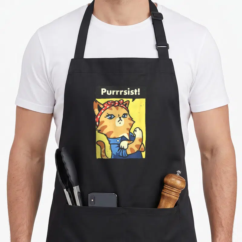 Tablier de cuisine noir avec illustration de chatte orange en overalls bleus et foulard à pois rouges sur fond jaune.