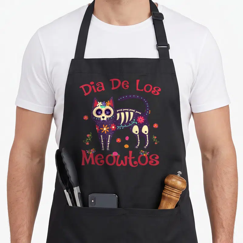 Tablier de cuisine noir avec un chat squelette violet orné de fleurs et du texte Dia De Los Meowtos en rouge.