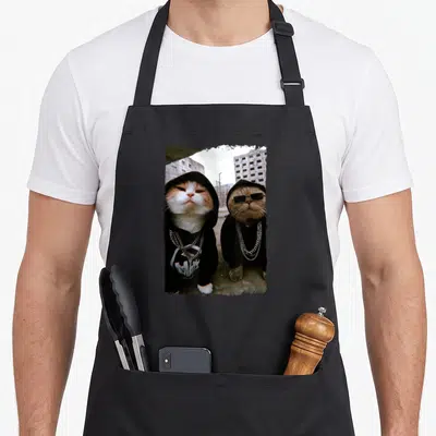 Tablier de cuisine noir avec deux chats en tenue streetwear portant des lunettes noires et des chaînes métalliques devant un décor urbain.