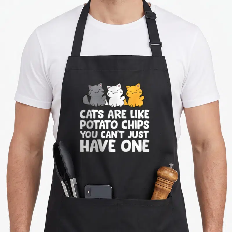 Tablier de cuisine noir avec trois chats colorés et une citation humoristique en anglais blanc.