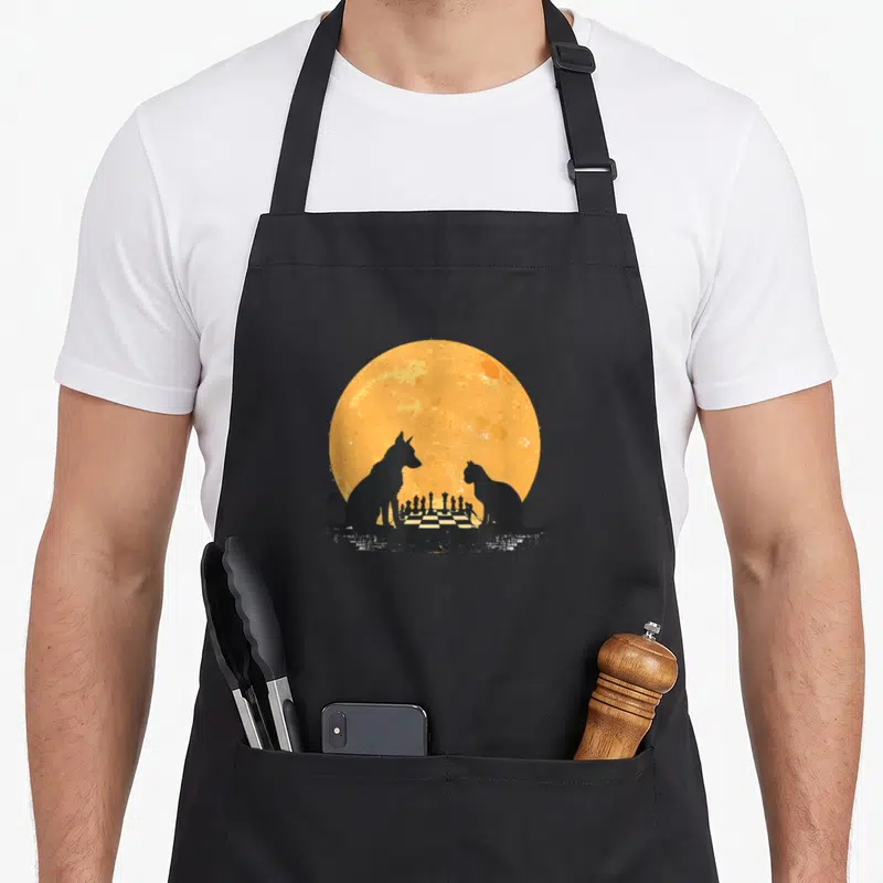 Tablier de cuisine noir avec silhouette chien et chat jouant aux échecs devant un disque orange texturé.