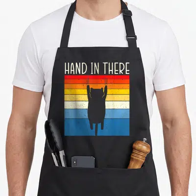 Tablier de cuisine noir avec silhouette de chat gris suspendu sur fond de bandes colorées horizontales et texte blanc en haut.