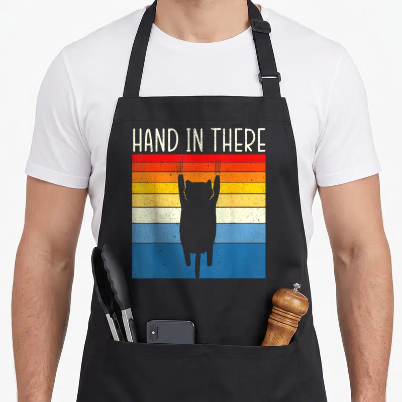 Tablier de cuisine noir avec silhouette de chat gris suspendu sur fond de bandes colorées horizontales et texte blanc en haut.