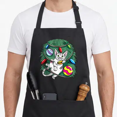 Tablier de cuisine noir avec un chaton blanc et gris intégré à un arbre de Noël vert orné de boules colorées et de lumières.