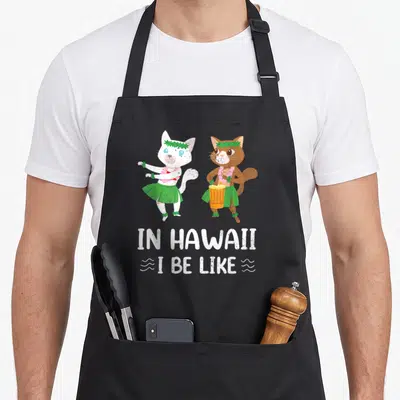 Tablier de cuisine noir avec deux chats en tenue hawaïenne et le texte In Hawaii I Be Like imprimé en blanc.