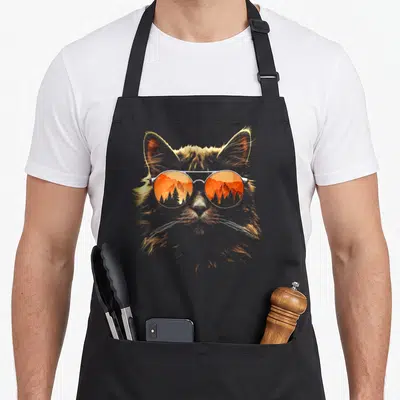 Tablier de cuisine noir avec tête de chat aux lunettes de soleil reflétant un paysage montagneux orange.