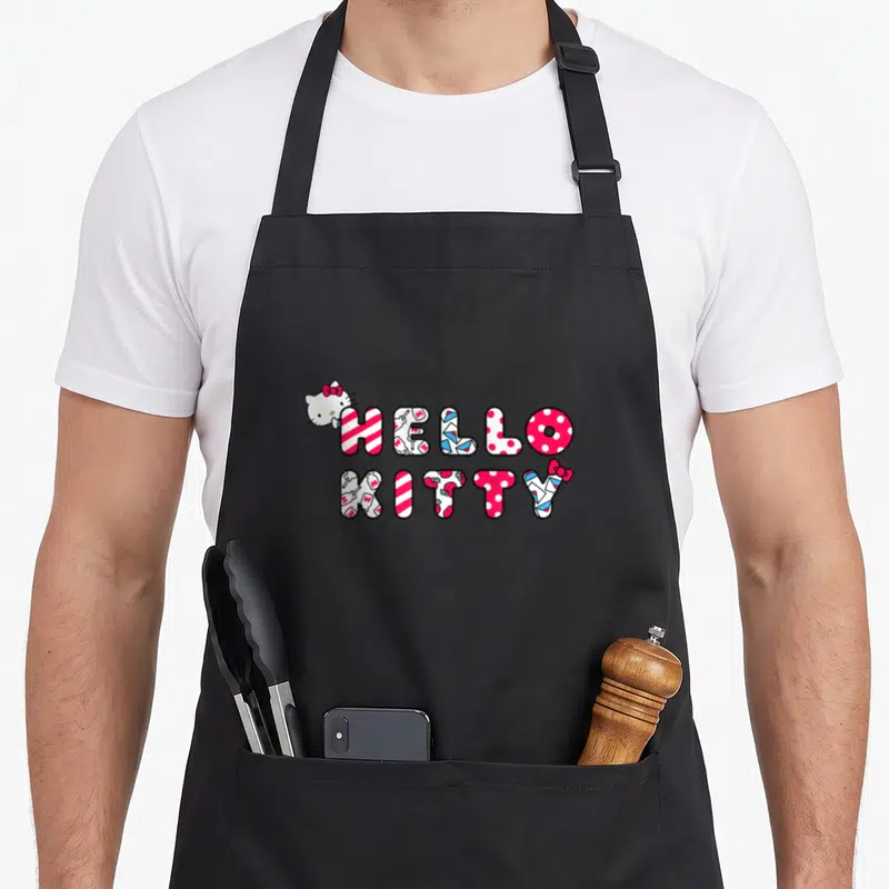 Tablier de cuisine noir avec le logo Hello Kitty en lettres colorées et un petit chat blanc à l'angle supérieur gauche.