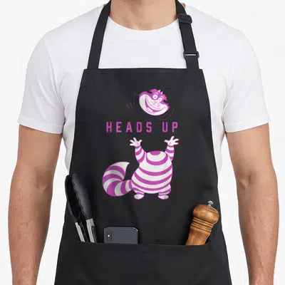 Tablier de cuisine noir avec illustration rose et blanche du Cheshire Cat et l'inscription Heads Up au centre.