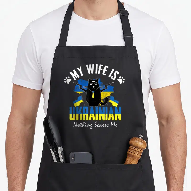 Tablier de cuisine noir avec illustration d'un chat noir en costume tenant deux pistolets devant un drapeau ukrainien.