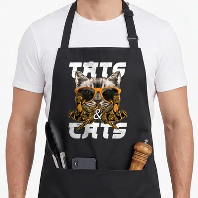 Tablier de cuisine noir avec tête de chat portant des lunettes orange et deux moteurs croisés en dessous.