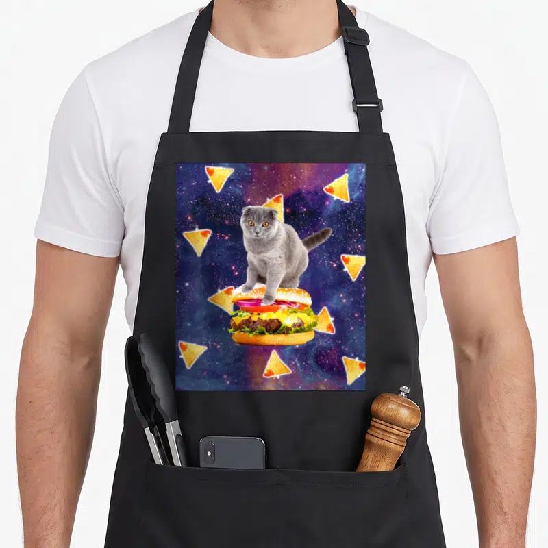 Tablier de cuisine noir avec un chat gris posant sur un burger géant entouré de nachos dans l'espace.