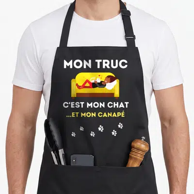 Tablier de cuisine noir avec une femme allongée sur un canapé jaune caressant un chat blanc entouré de traces de pattes.