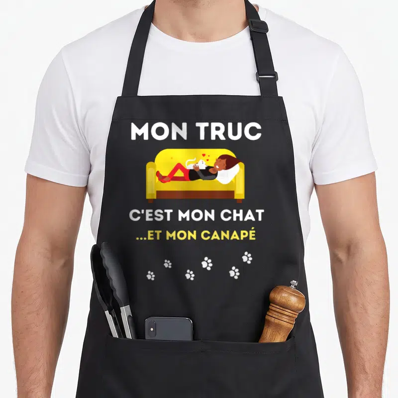 Tablier de cuisine noir avec une femme allongée sur un canapé jaune caressant un chat blanc entouré de traces de pattes.