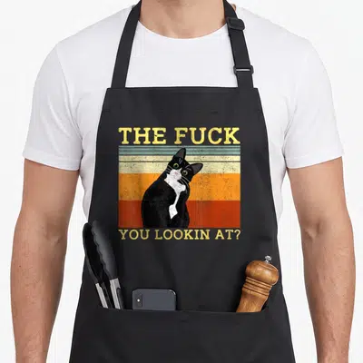 Tablier de cuisine noir avec illustration vintage d'un chat bicolore aux yeux verts et texte jaune provocateur.