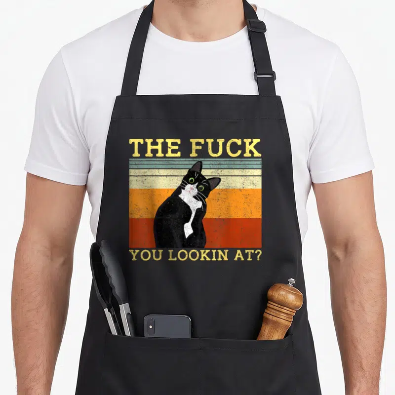 Tablier de cuisine noir avec illustration vintage d'un chat bicolore aux yeux verts et texte jaune provocateur.